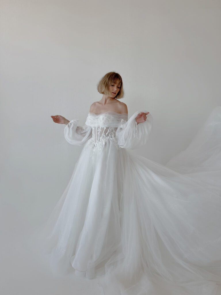 Bride wearing a strapless lace wedding gown with matching sheer opera gloves beside a sunlit window.