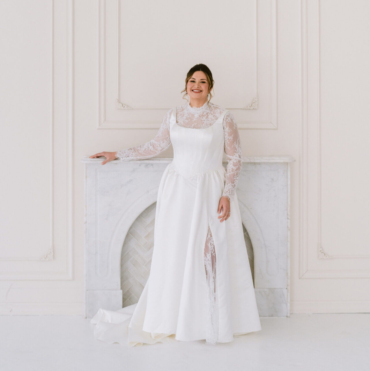 Bride wearing a long-sleeve minimalist wedding gown standing in an elegant white room with classic wall paneling.