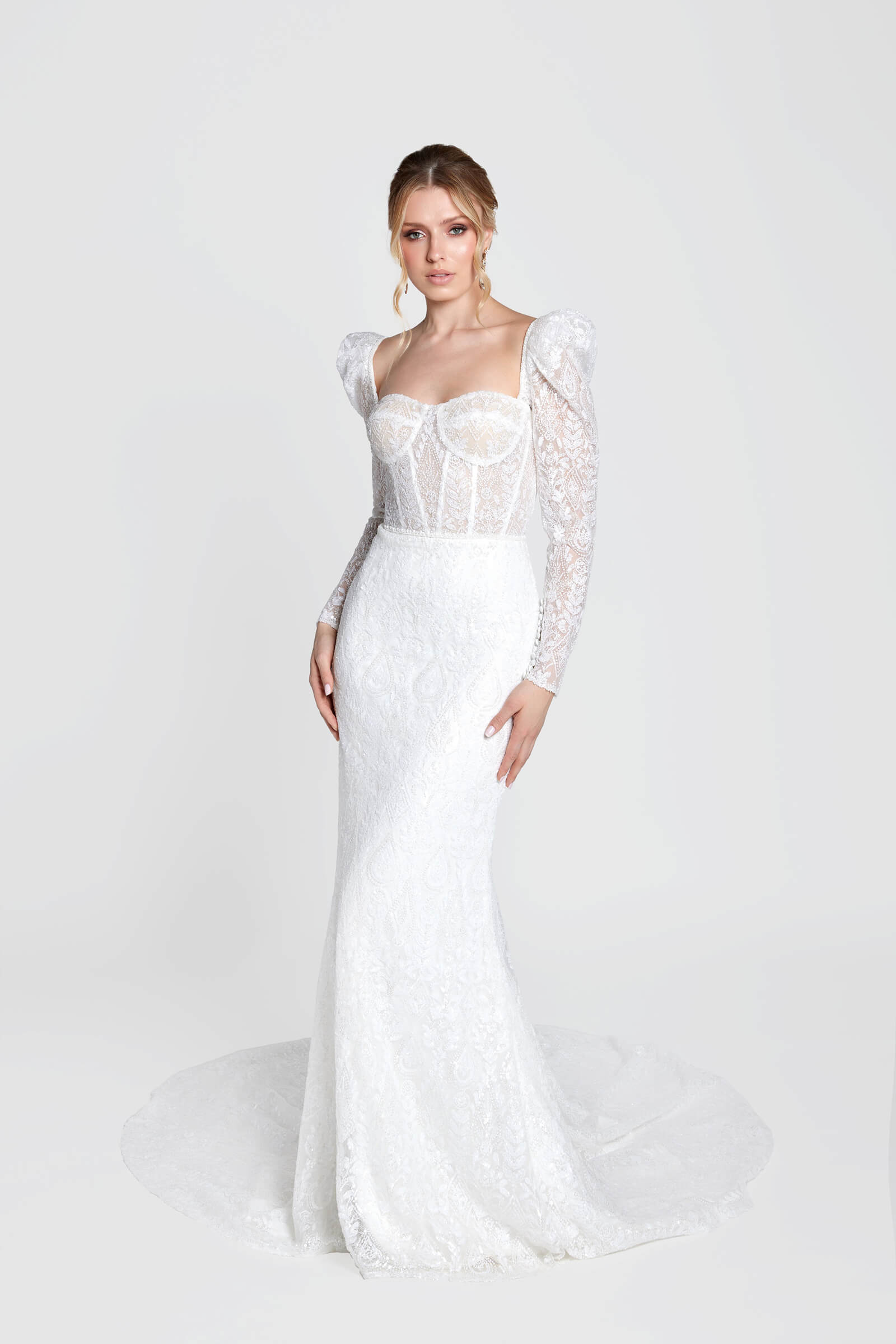 Bride wearing a fitted lace mermaid wedding gown with long illusion sleeves in a bright studio.