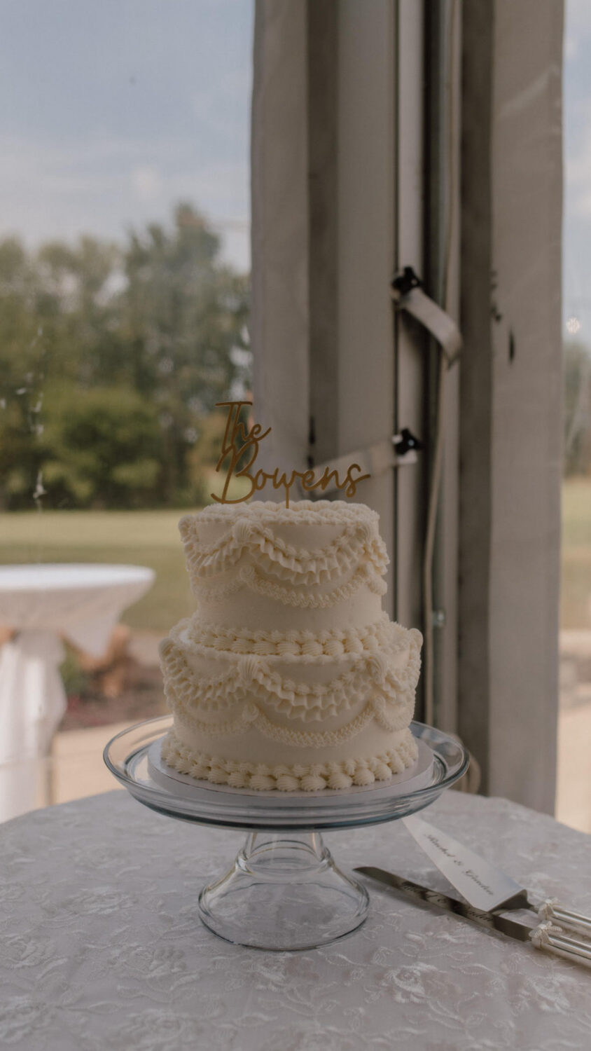 A two-tier white wedding cake with piped buttercream details and a custom cake topper sits on a glass pedestal near a window overlooking an outdoor landscape.