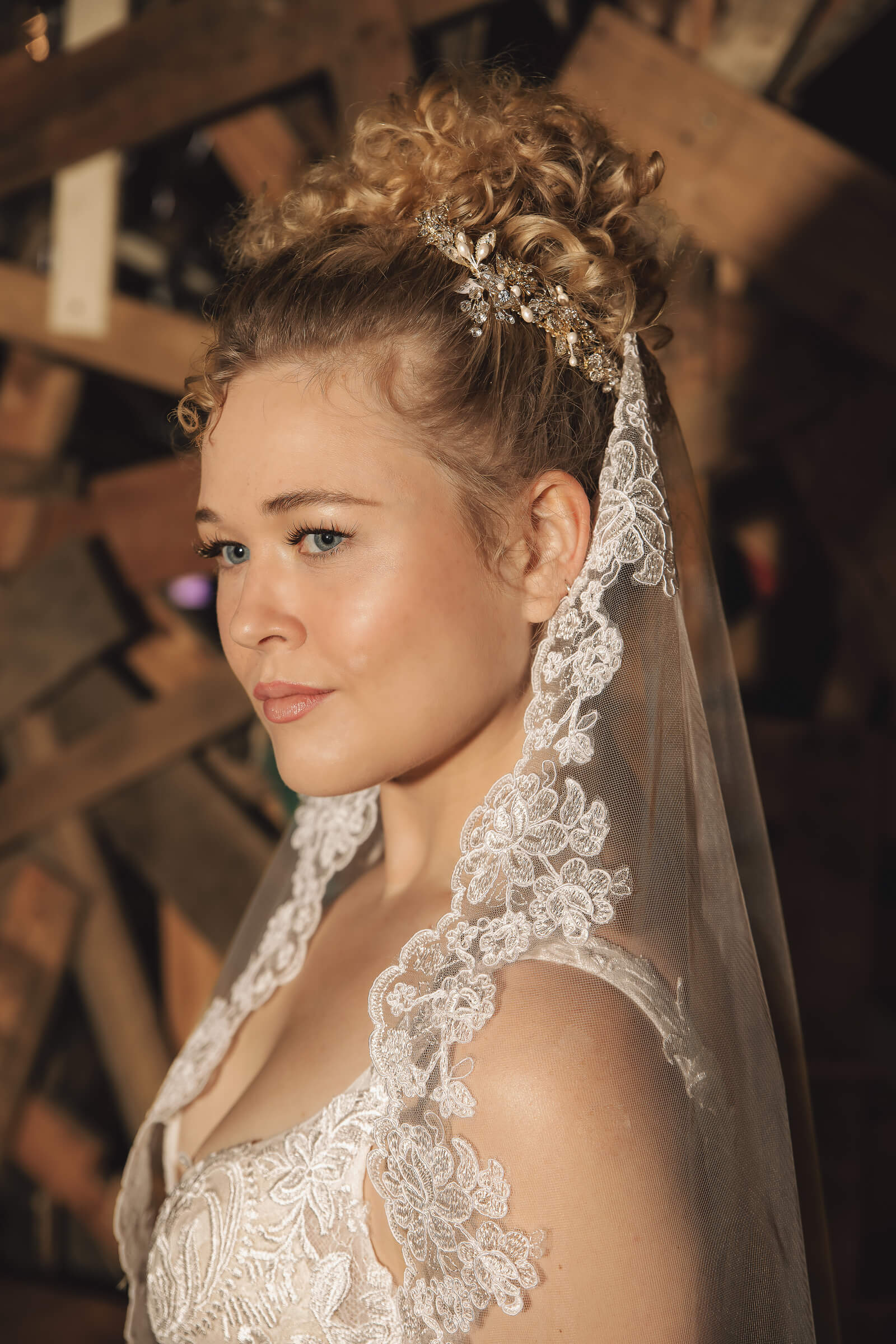 A bride with a curly updo wears a jeweled pearl hairpiece and a veil edged with intricate floral lace.