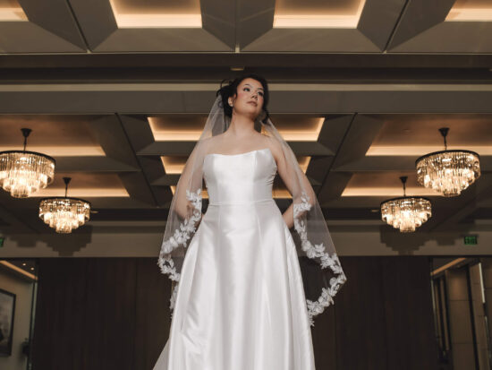 A bride in a satin wedding gown poses indoors beneath chandeliers while wearing a veil with floral lace appliqués.