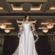 A bride in a satin wedding gown poses indoors beneath chandeliers while wearing a veil with floral lace appliqués.