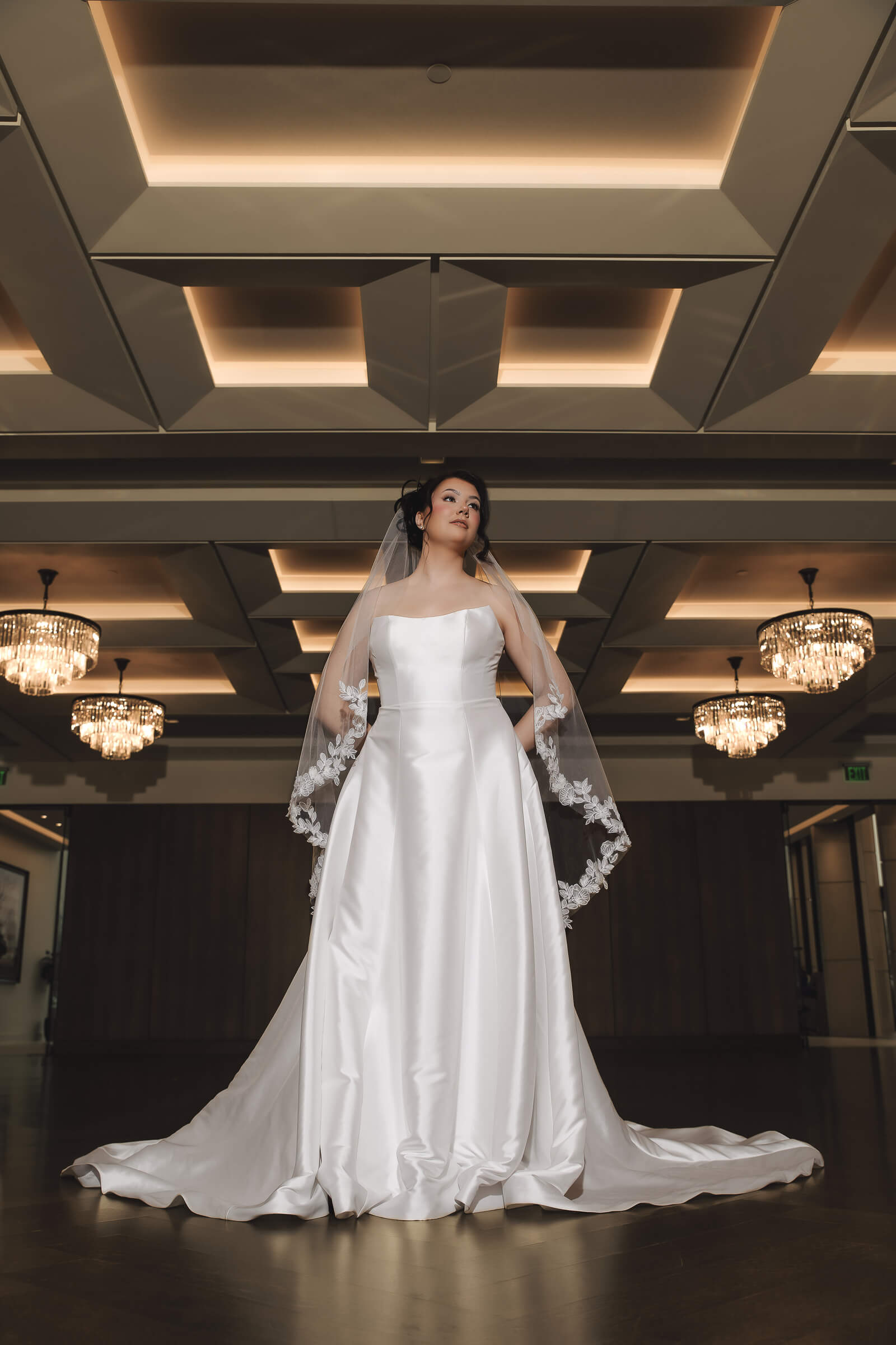 A bride in a satin wedding gown poses indoors beneath chandeliers while wearing a veil with floral lace appliqués.