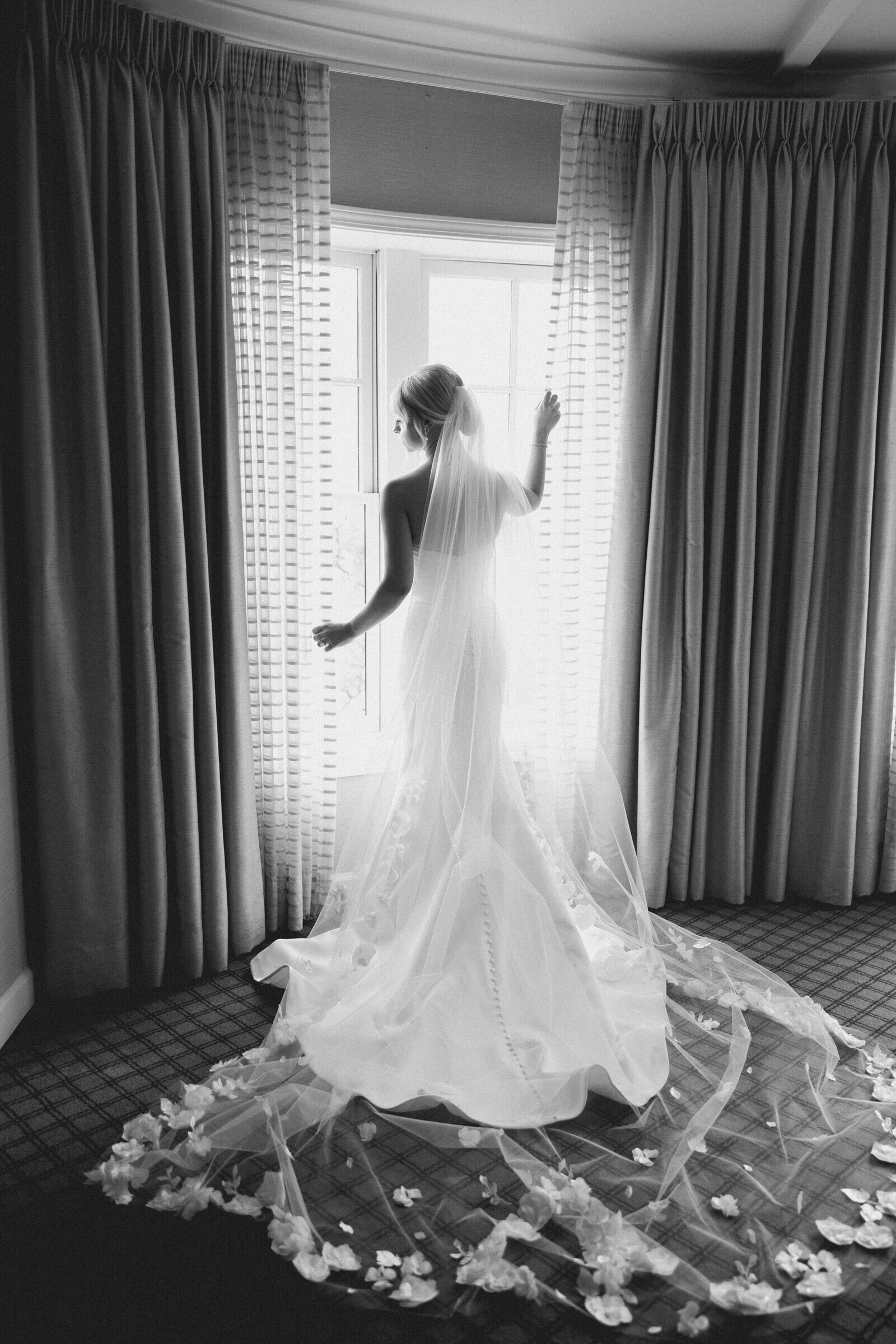 A bride in a white gown stands by a window, highlighting her elegant hairstyle and bridal accessories.