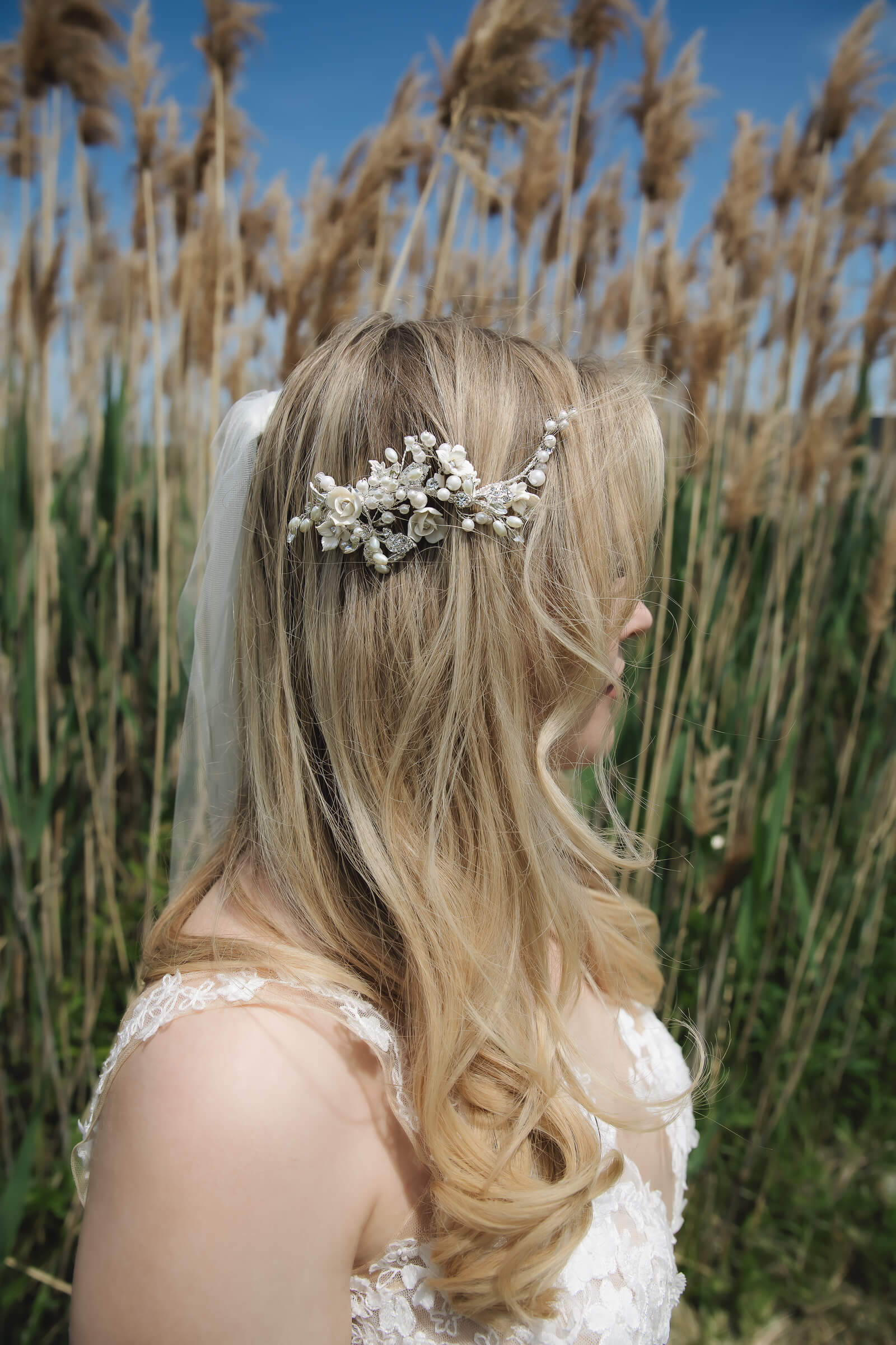 A bride with long blonde curls wears a floral and pearl hairpiece while standing outdoors among tall grasses.