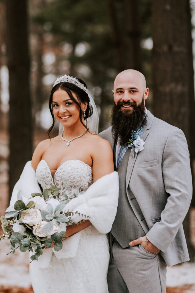 A bride and groom pose together outdoors, with the bride holding a bouquet and wearing a white wedding gown.
