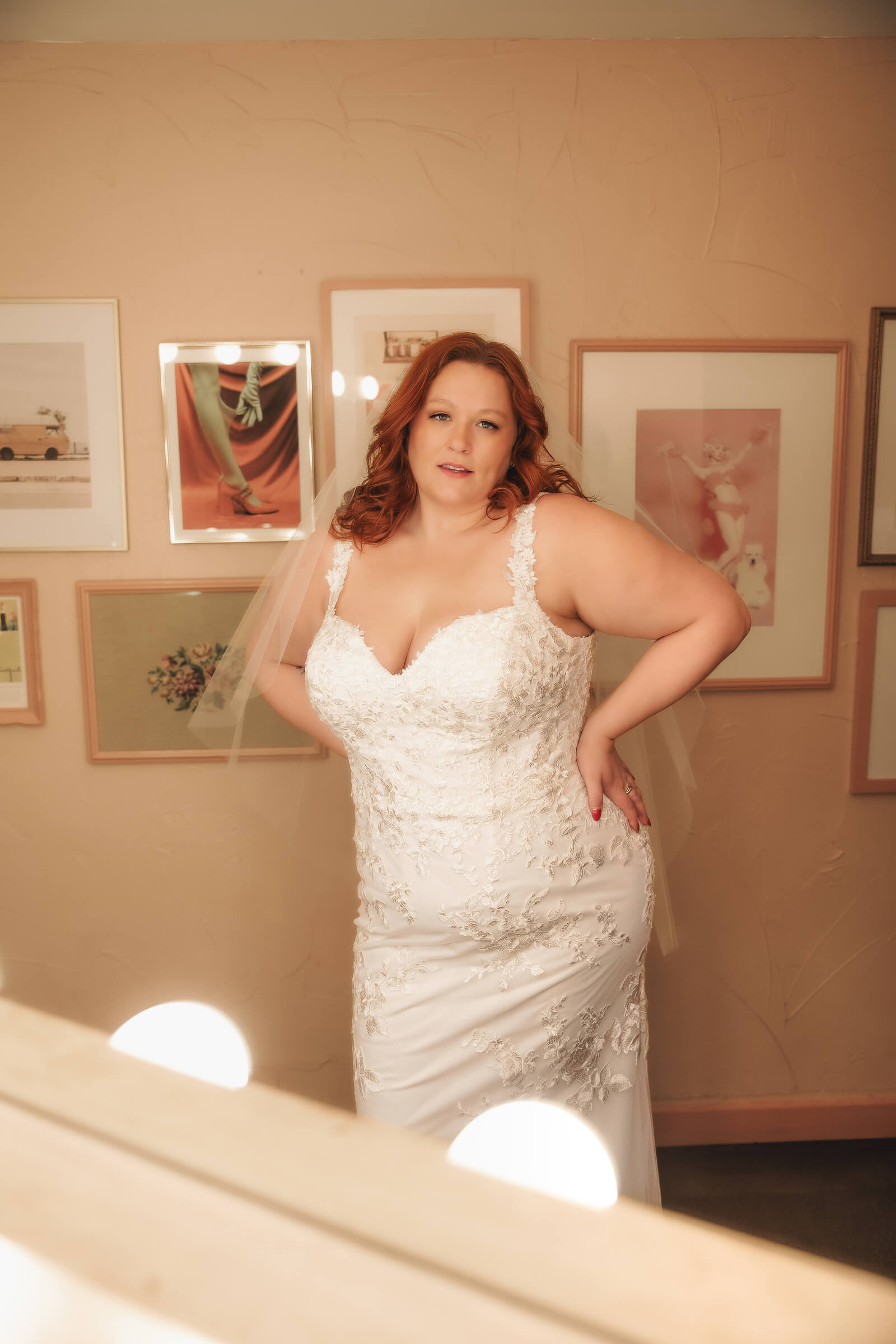 A bride with red hair poses in a fitted lace wedding dress and sheer veil in front of a gallery wall.