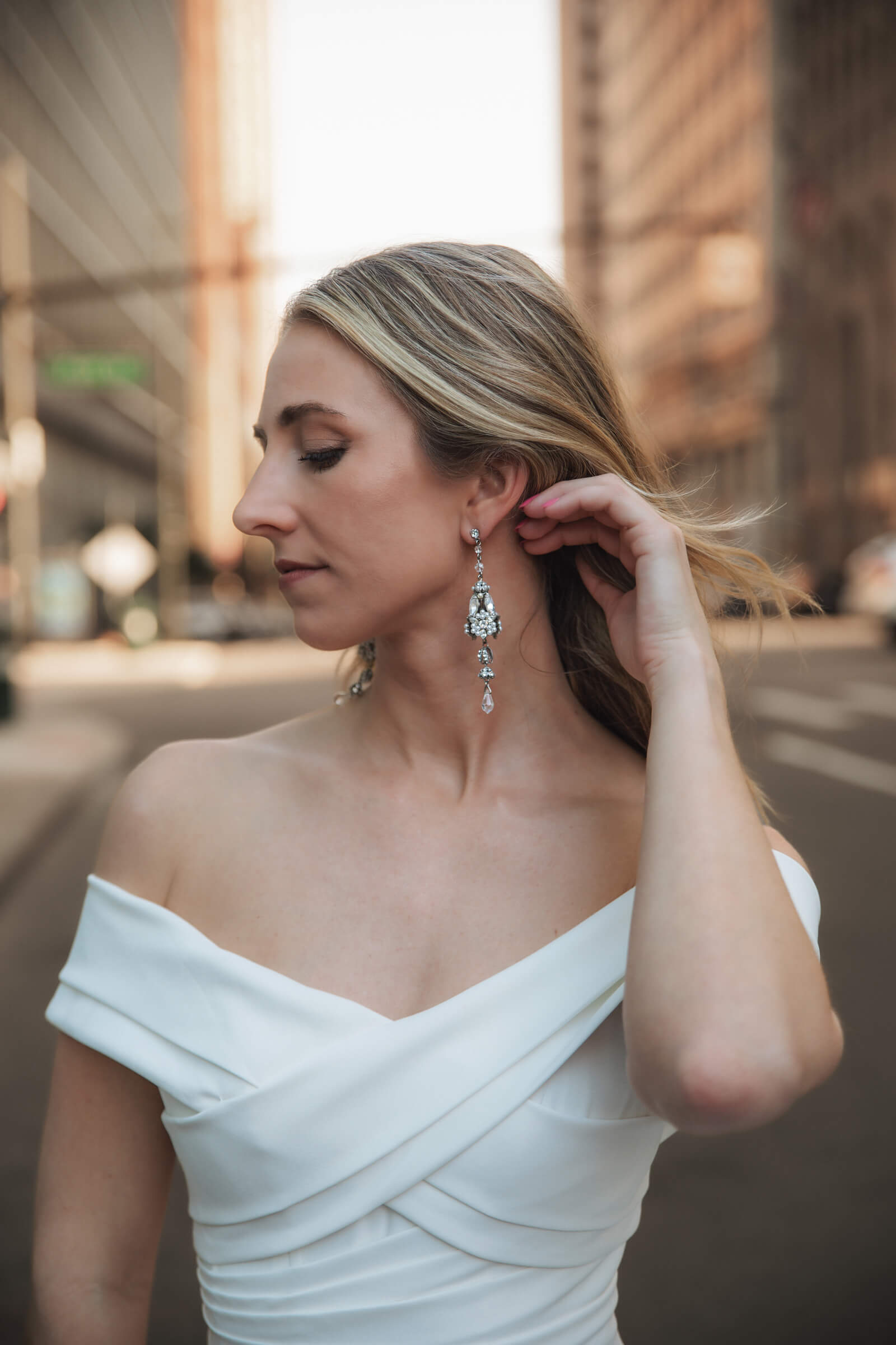 A bride in an off-the-shoulder wedding gown adjusts her earring, highlighting her hairstyle and bridal jewelry.
