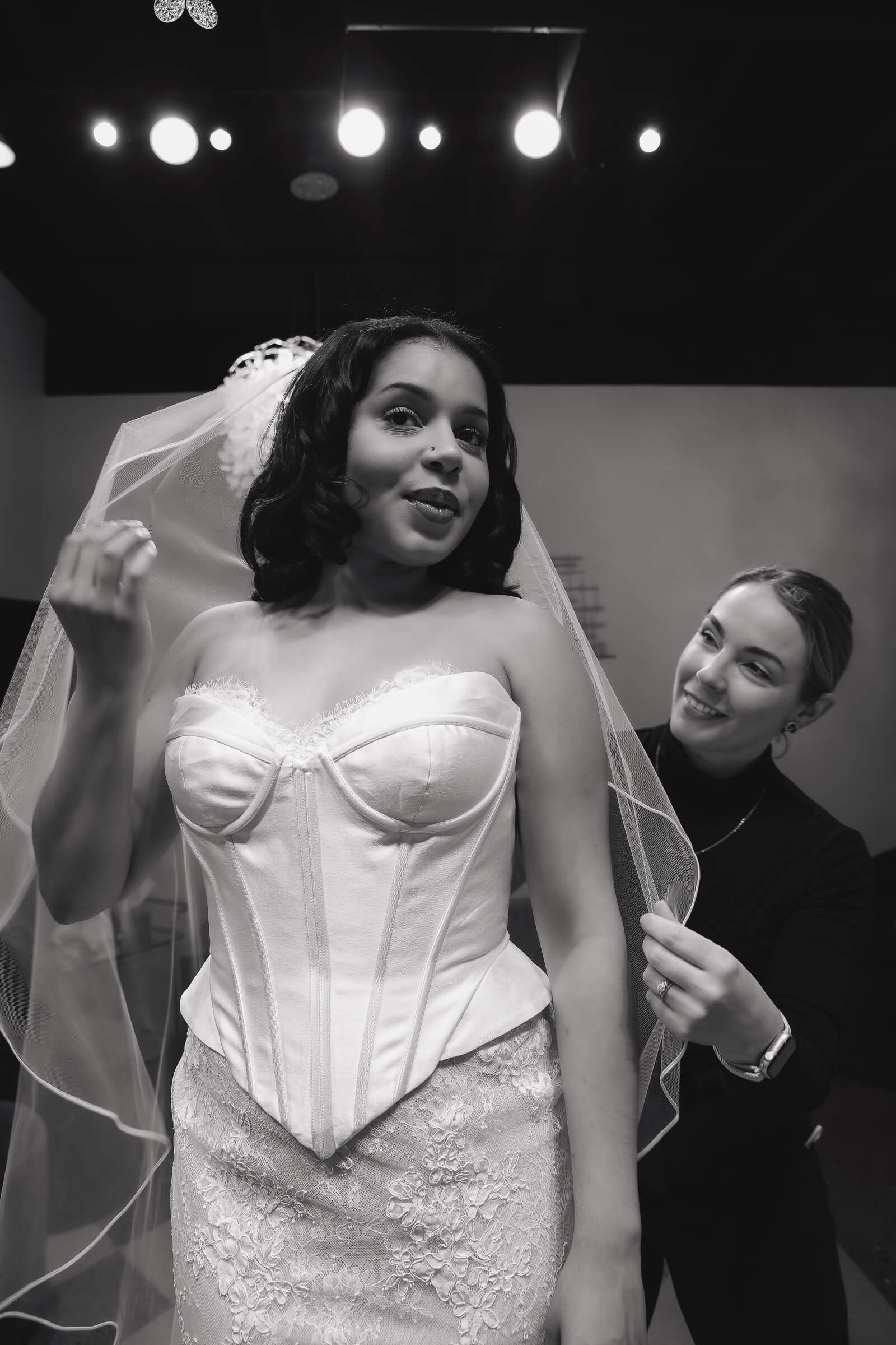 A bride in a white wedding gown poses backstage while another woman helps with her hairstyle and accessories.