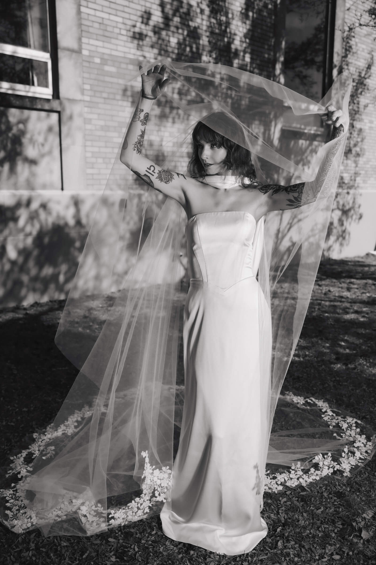 A bride in a strapless wedding gown holds a long sheer veil above her head in a black-and-white outdoor portrait.