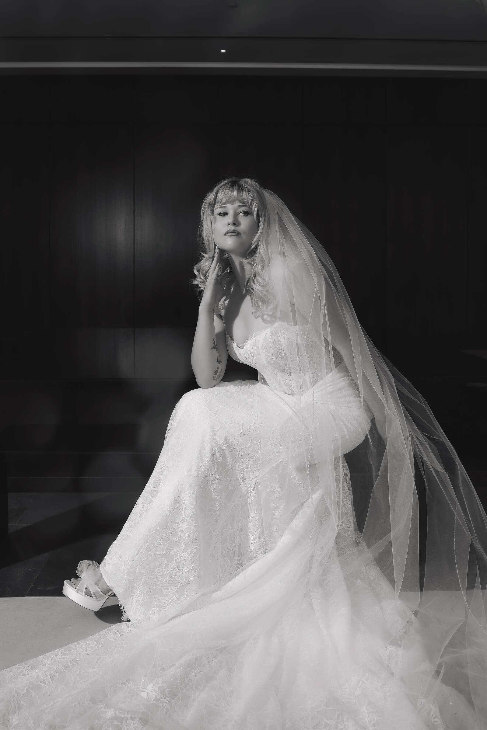 A bride in a lace wedding gown and long sheer veil sits in dramatic lighting in a black-and-white portrait.