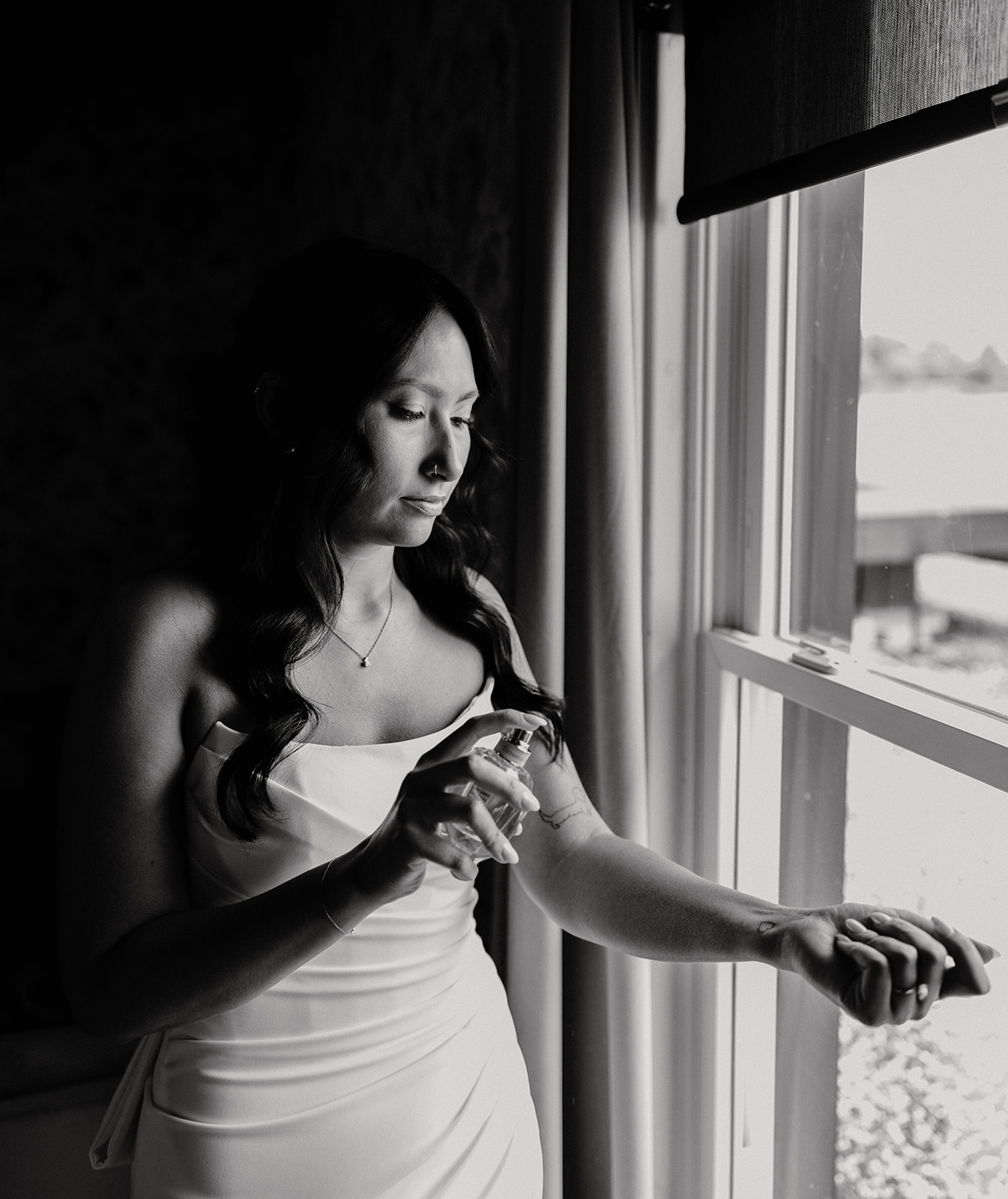 A bride in a white wedding gown adjusts her hair beside a window, showcasing her bridal hairstyle.