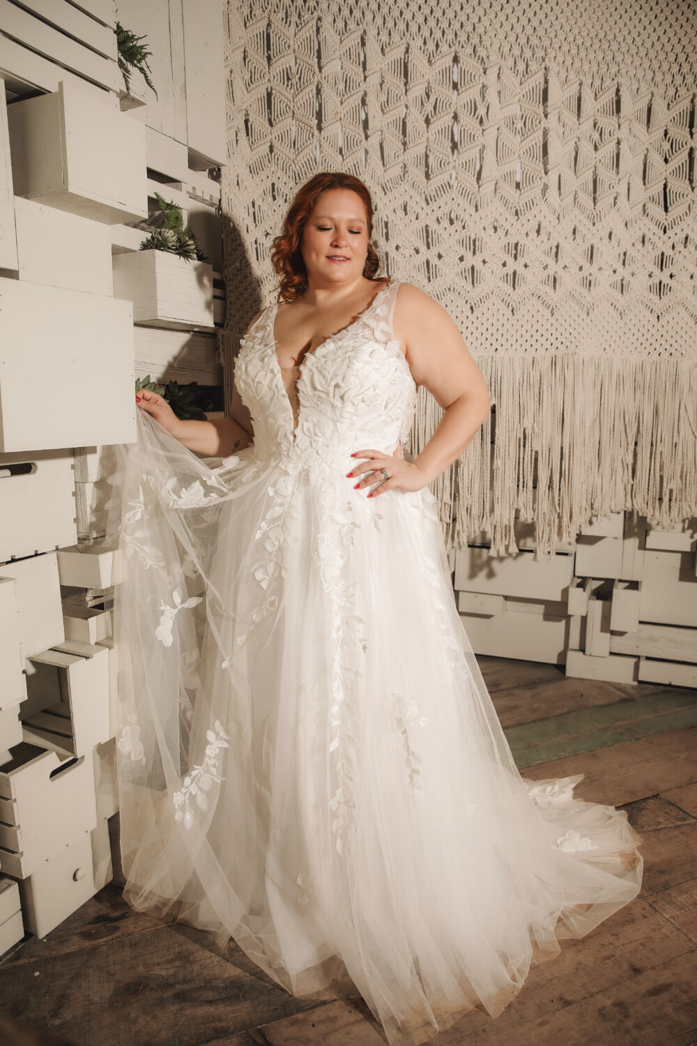 A woman poses in a white lace wedding dress with a plunging neckline, floral appliqués, and a flowing tulle skirt in a sunlit room.