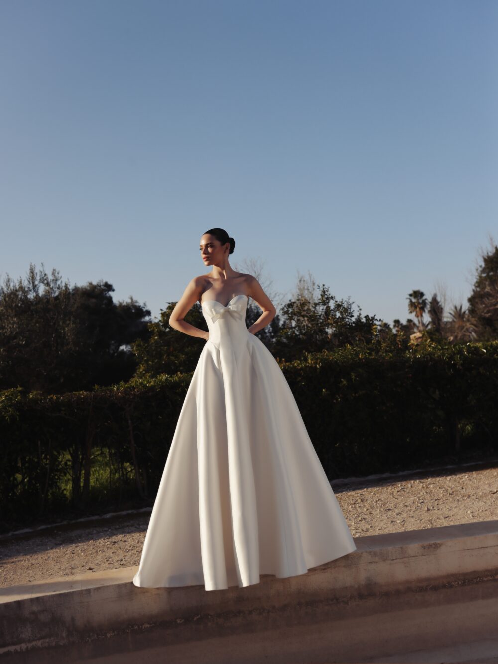 A woman stands outdoors in a strapless white ball gown with a bow detail at the neckline and a full structured skirt.