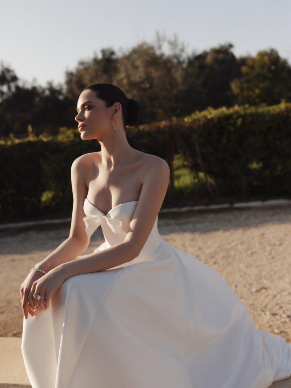 A woman sits outdoors in a strapless white wedding gown with a bow at the neckline and a voluminous skirt.