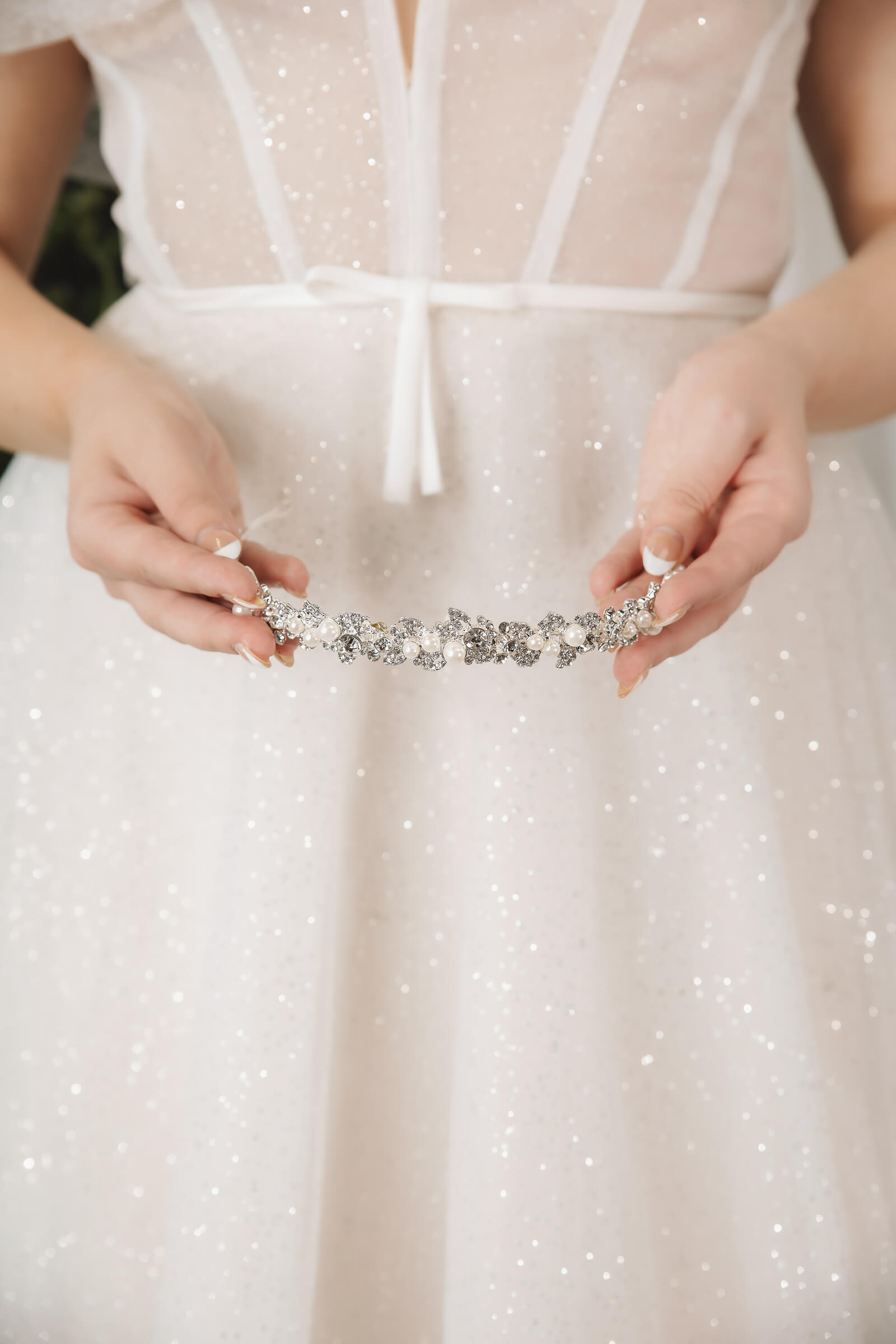 A bride holds a silver-toned hair accessory decorated with crystals and pearls against her sparkling white gown.