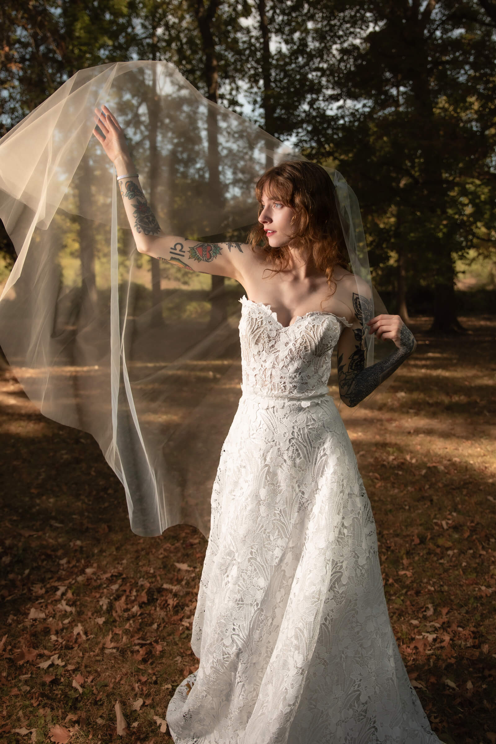 A bride in a white wedding gown poses outdoors while lifting her veil, showcasing her bridal hairstyle and accessories.