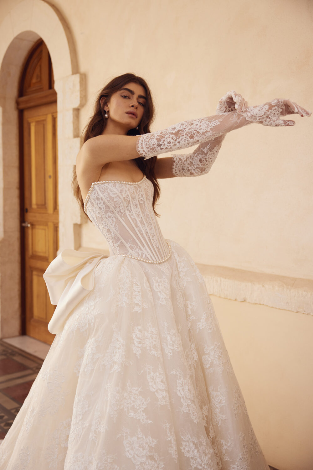 A woman wears a lace corset wedding gown with pearl trim, a full lace skirt, long lace gloves, and a large bow at the back.