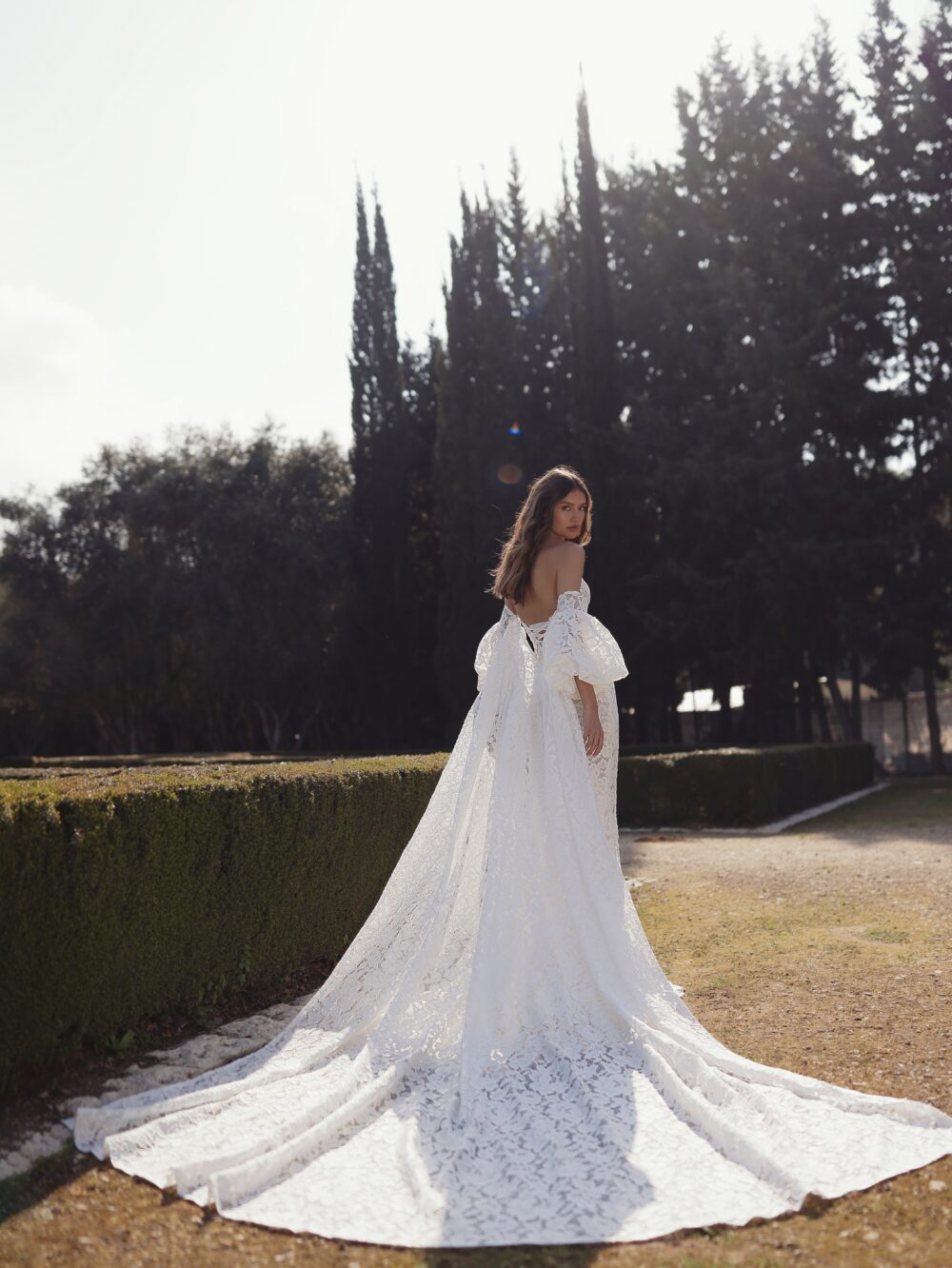 A woman looks over her shoulder while wearing an off-the-shoulder lace wedding gown with voluminous sleeves and an exceptionally long train.