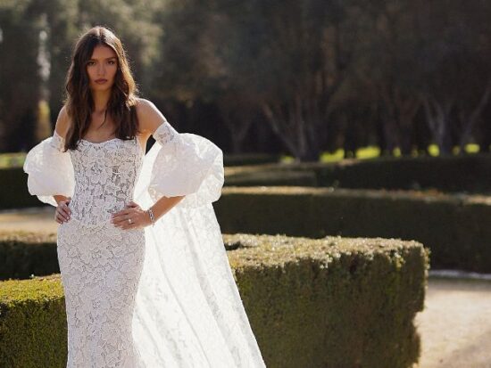 A woman wears a fitted lace wedding gown with dramatic puff sleeves and a flowing lace overskirt while posing in a manicured garden.
