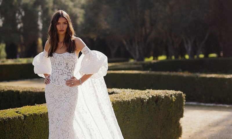 A woman wears a fitted lace wedding gown with dramatic puff sleeves and a flowing lace overskirt while posing in a manicured garden.