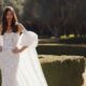 A woman wears a fitted lace wedding gown with dramatic puff sleeves and a flowing lace overskirt while posing in a manicured garden.