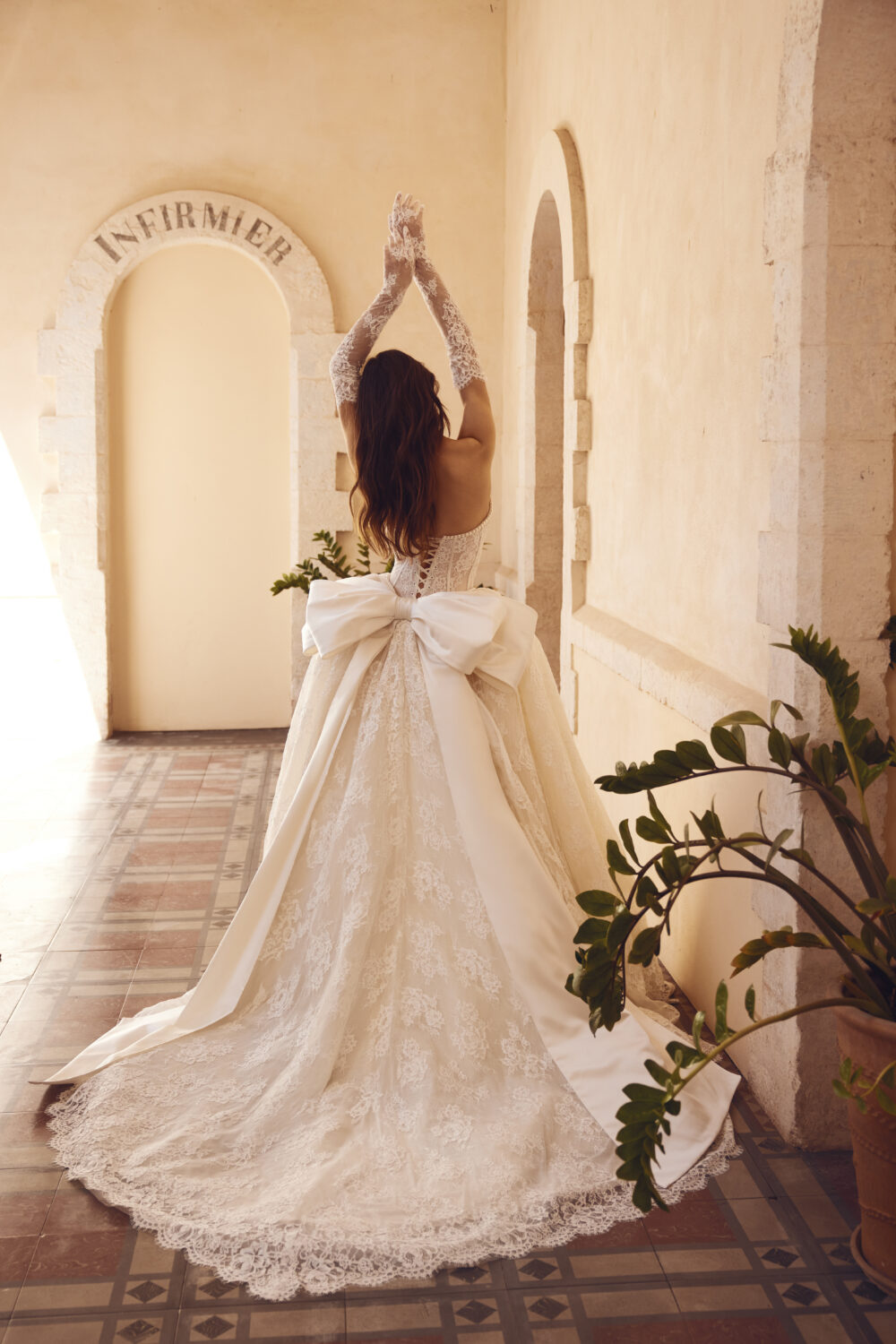 A woman poses from behind in an ornate lace wedding gown with long lace gloves, a corset back, an oversized bow, and a sweeping train.
