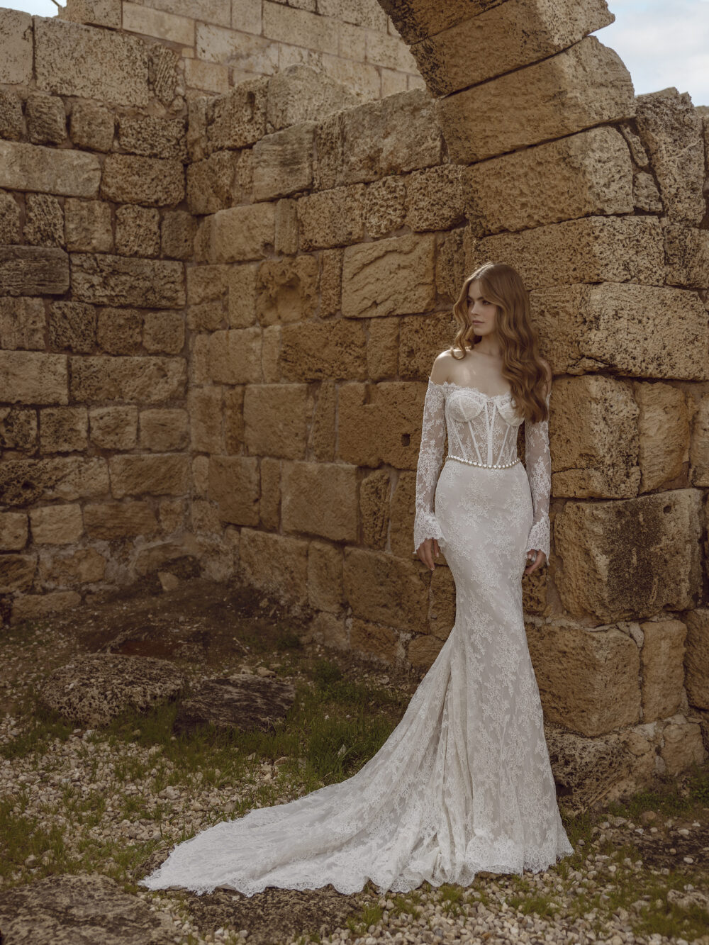 A woman stands among stone ruins wearing a fitted off-the-shoulder lace wedding gown with sheer long sleeves and a flared train.