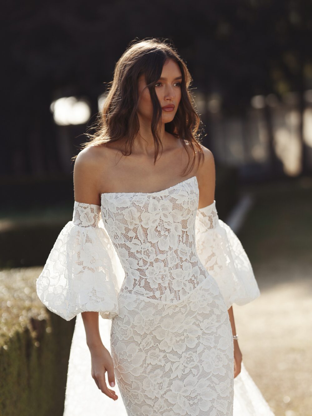 A woman poses outdoors in a fitted floral lace wedding gown with dramatic off-the-shoulder puff sleeves.