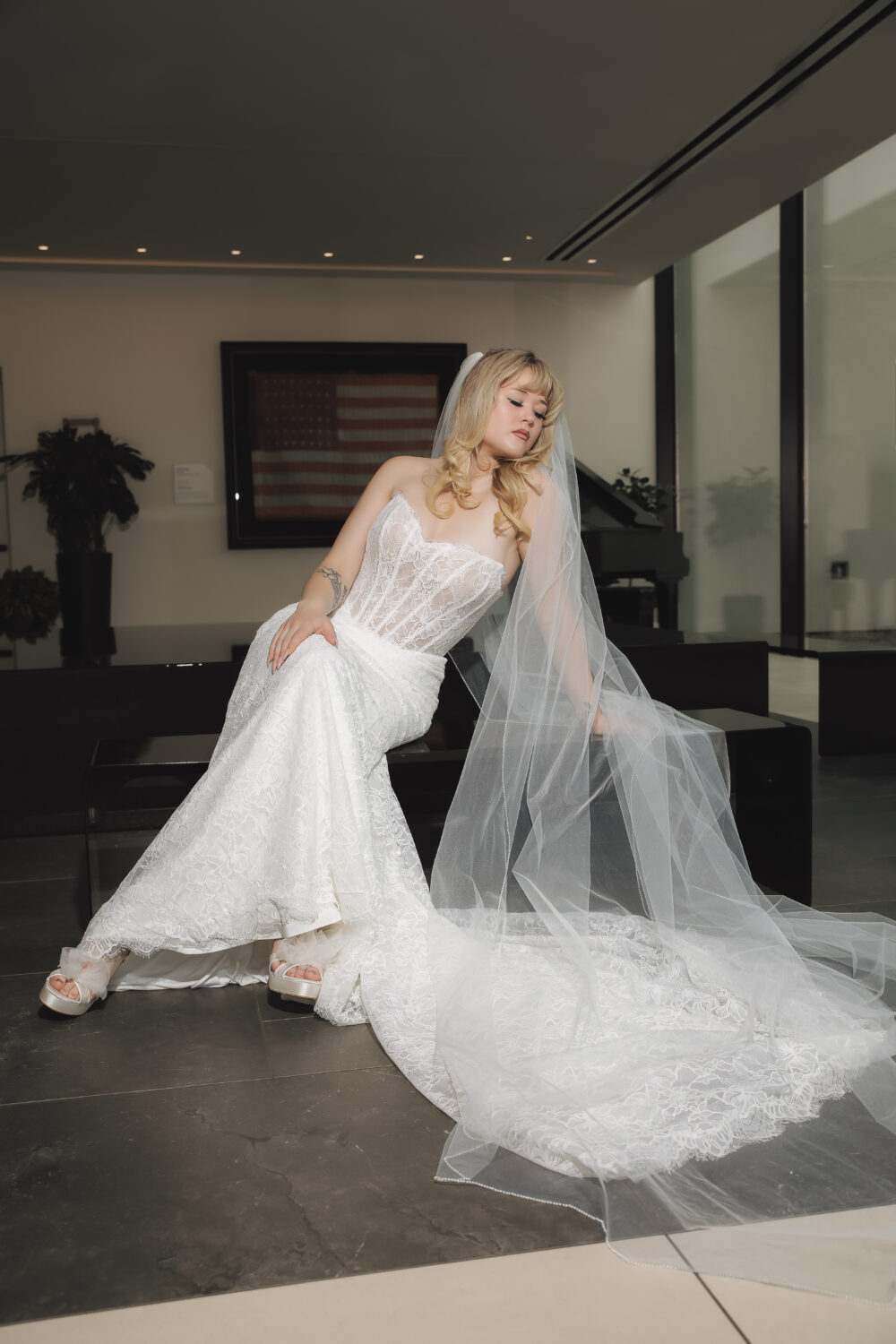 A woman sits in a strapless white lace wedding gown with a fitted silhouette, long train, and sheer veil draped across the floor.
