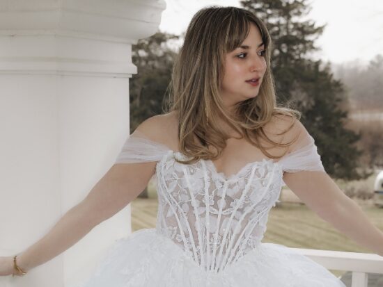 Bride wearing an off-the-shoulder wedding gown with a sheer lace corset bodice and voluminous layered tulle skirt on a porch.