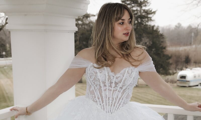 Bride wearing an off-the-shoulder wedding gown with a sheer lace corset bodice and voluminous layered tulle skirt on a porch.