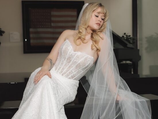 A woman in a strapless lace wedding gown sits beneath a long sheer veil in a modern interior with dark seating and large windows.