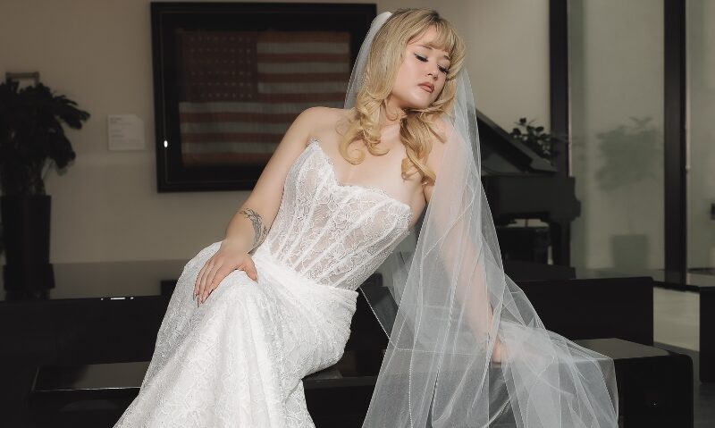 A woman in a strapless lace wedding gown sits beneath a long sheer veil in a modern interior with dark seating and large windows.