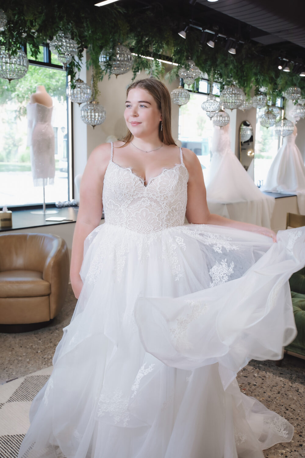 Bride in a lace wedding dress with thin straps holding out the layered tulle skirt while posing in a bridal boutique.