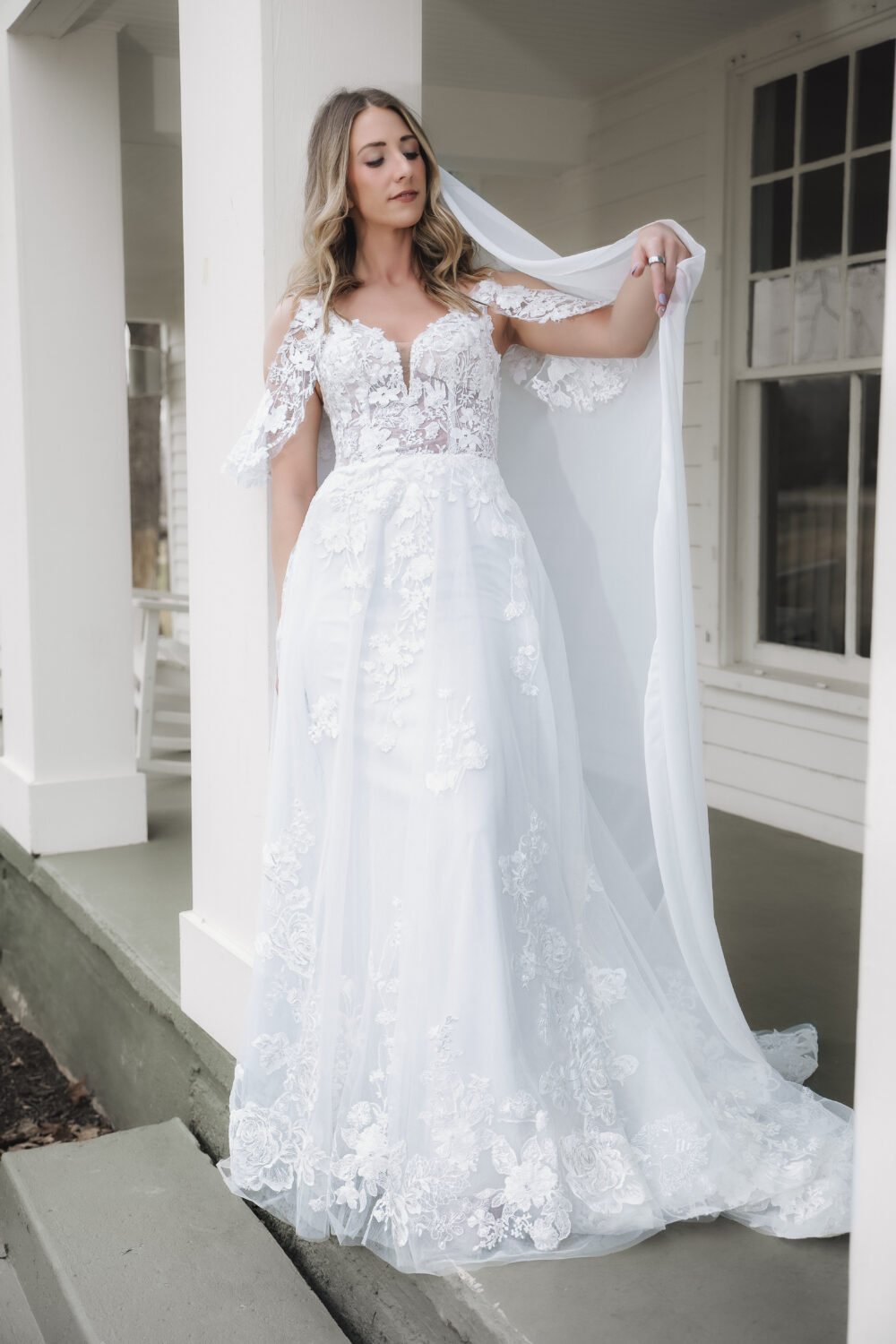 Bride posing on a porch in a lace wedding gown with floral appliqués, a flowing skirt, and a long veil.