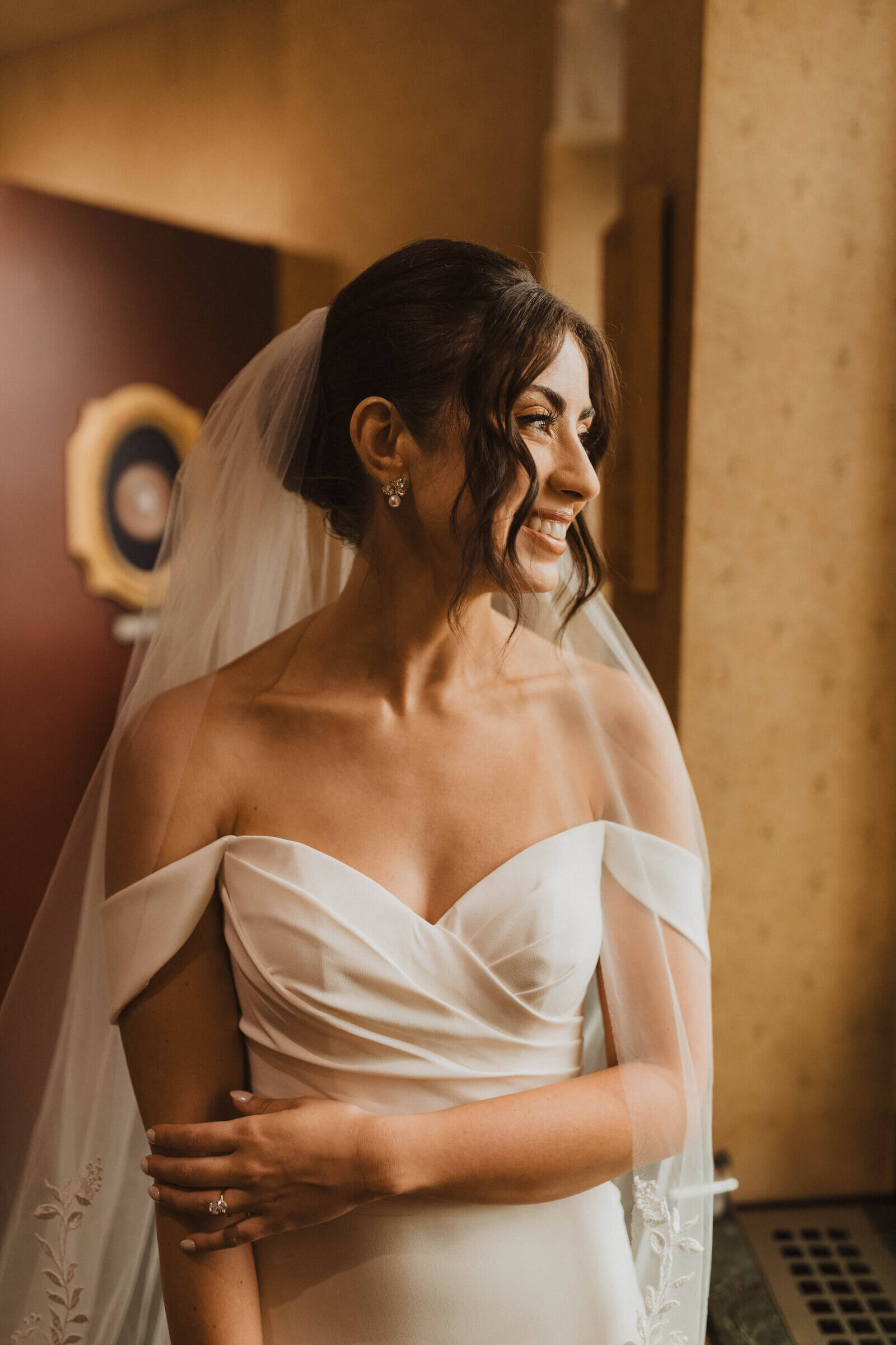 A bride in an off-the-shoulder wedding gown poses indoors, showcasing her updo hairstyle and earrings.