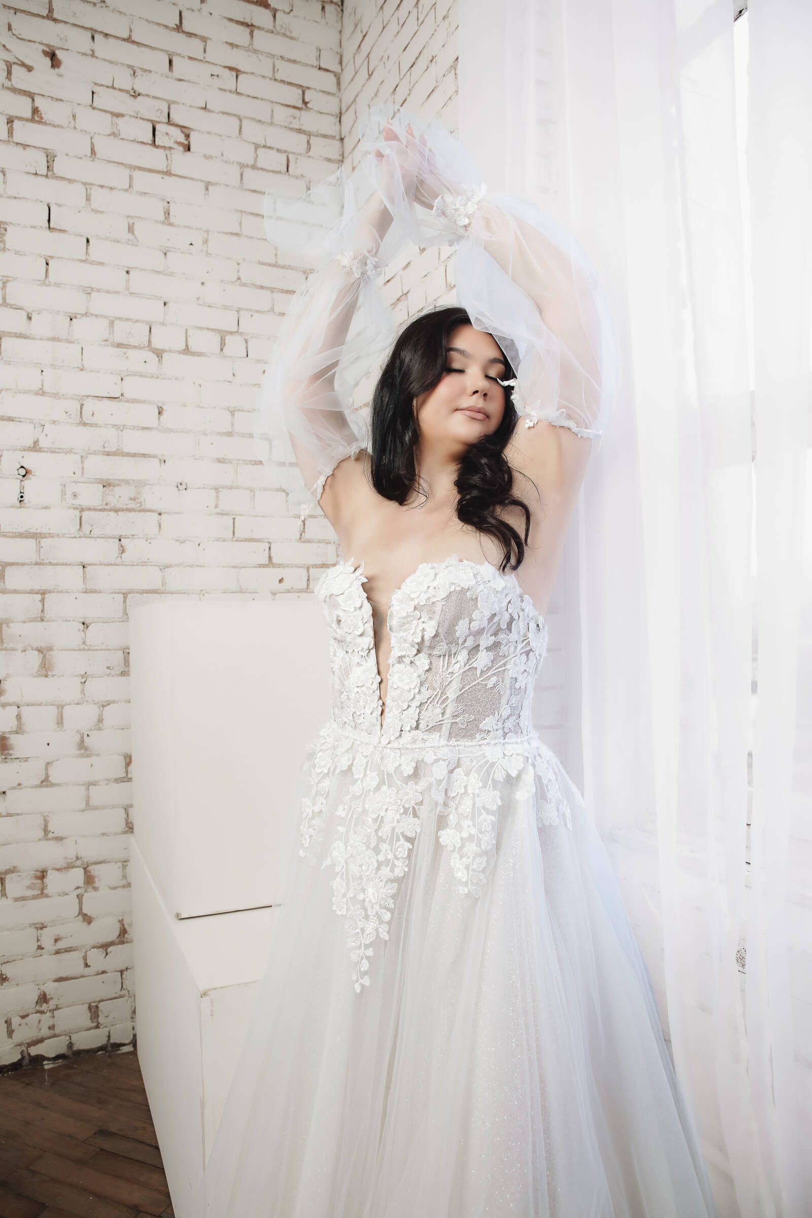 A bride in a floral lace wedding gown raises her arms while wearing sheer tulle gloves with floral details.