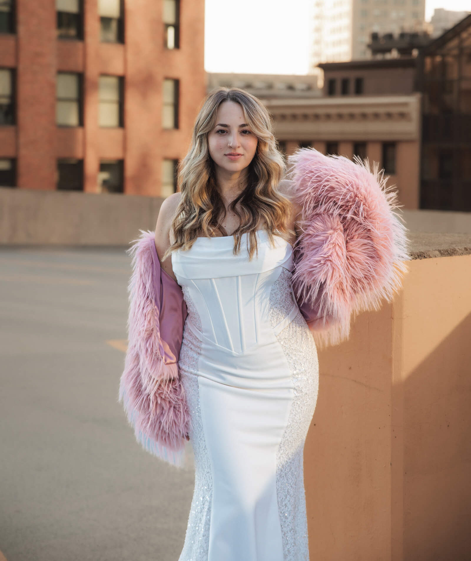 A bride in a white wedding gown and pink wrap poses on a rooftop, showcasing her hairstyle and bridal accessories.