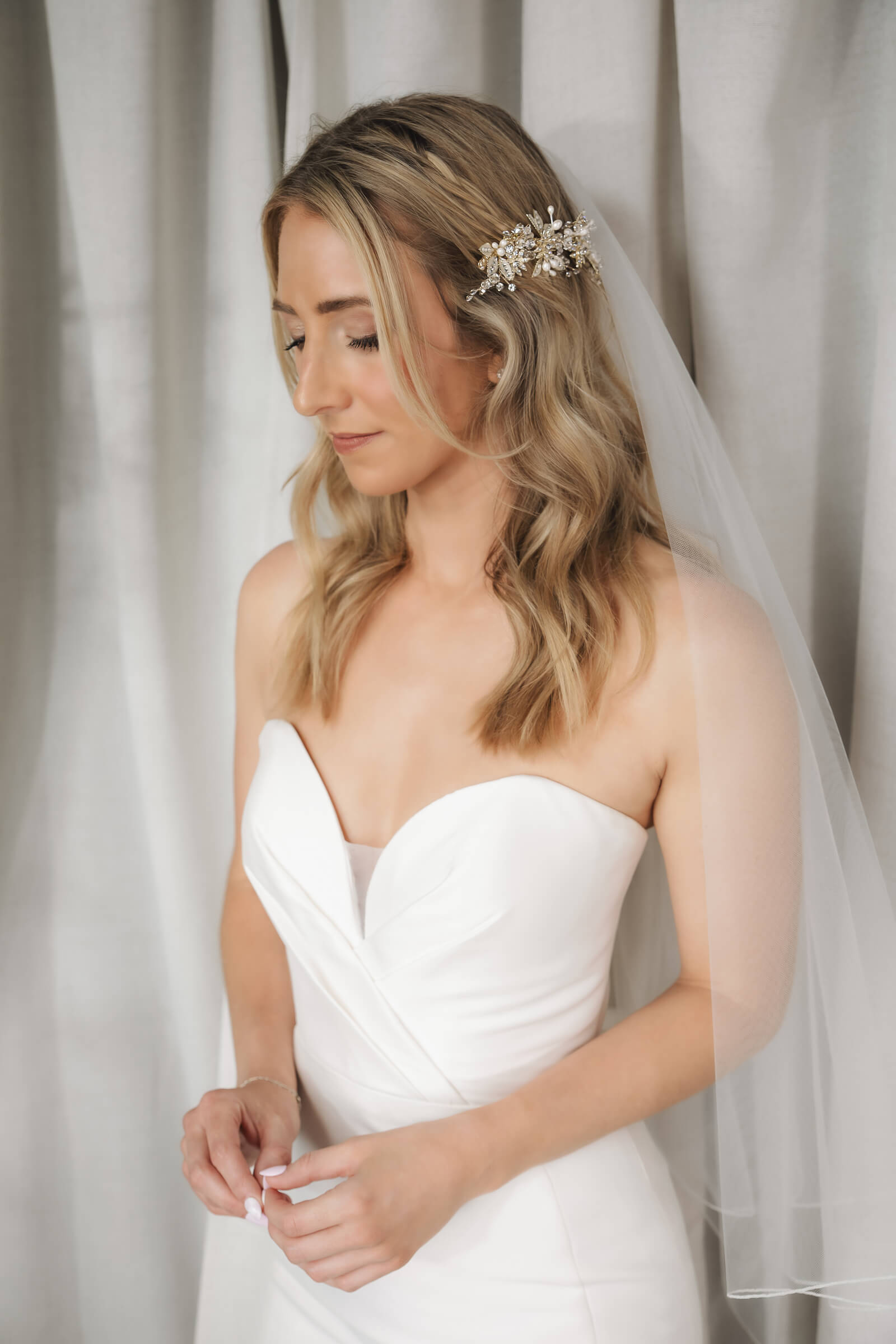 A bride with wavy blonde hair wears a jeweled floral hairpiece, sheer veil, and strapless white wedding gown.