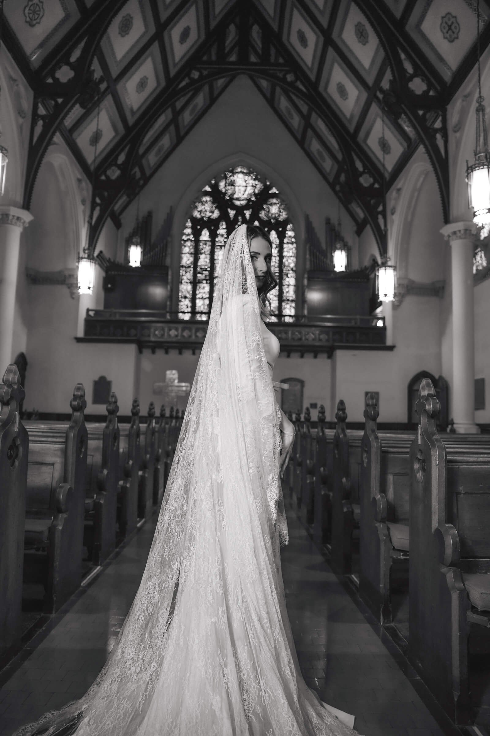A bride wearing a long lace veil stands between wooden pews in a church in a black-and-white portrait.