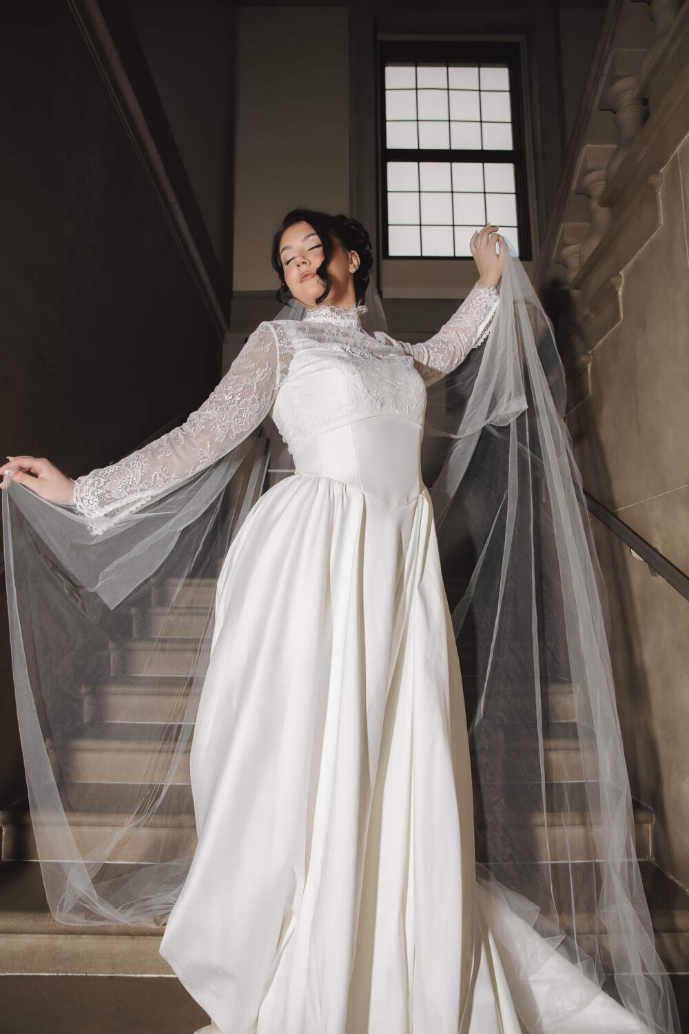 A bride in a white wedding gown stands behind a sheer veil, displaying her hairstyle and bridal accessories.
