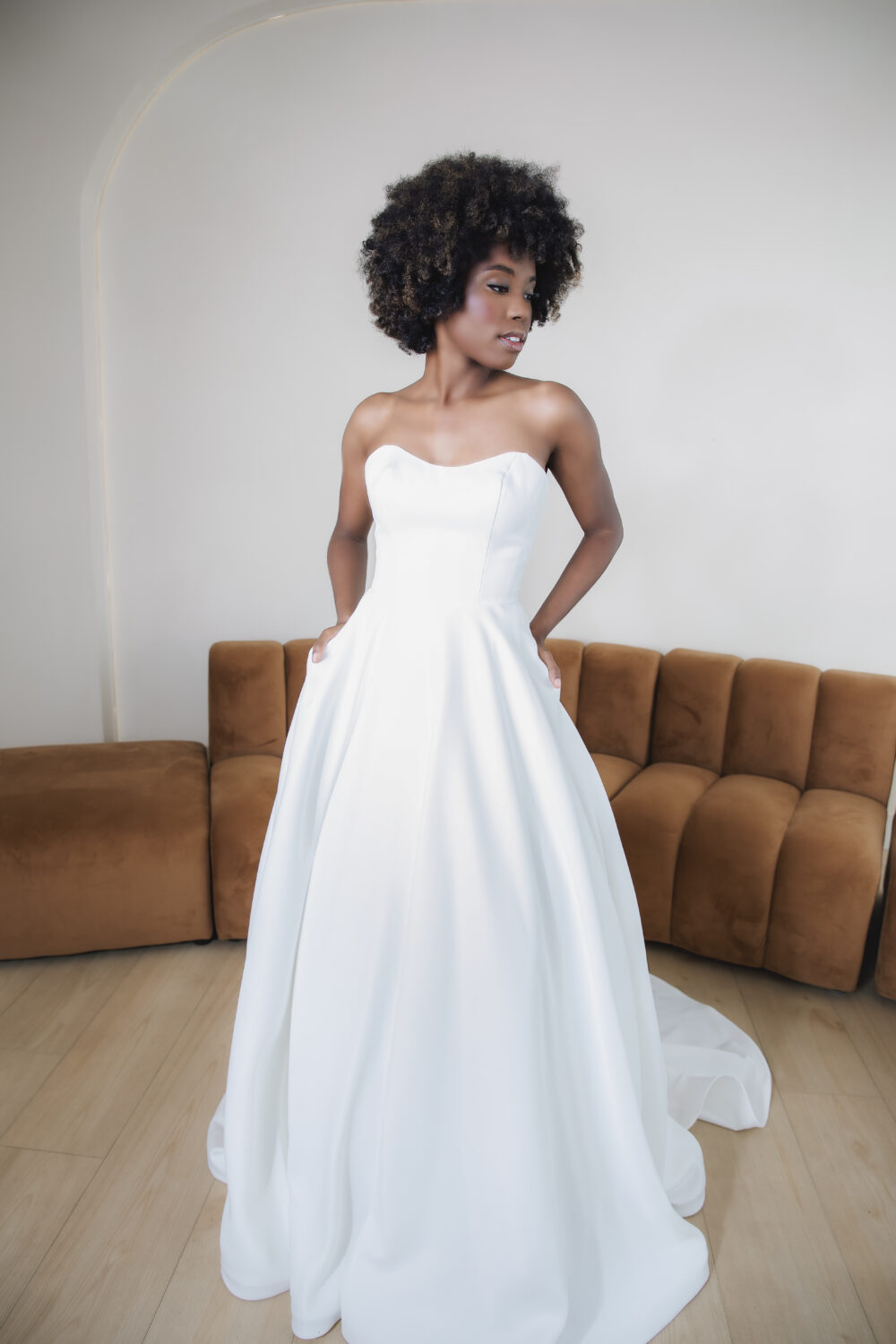 A woman poses in a simple strapless white ball gown with a sweetheart neckline and full satin skirt in front of a brown sofa.