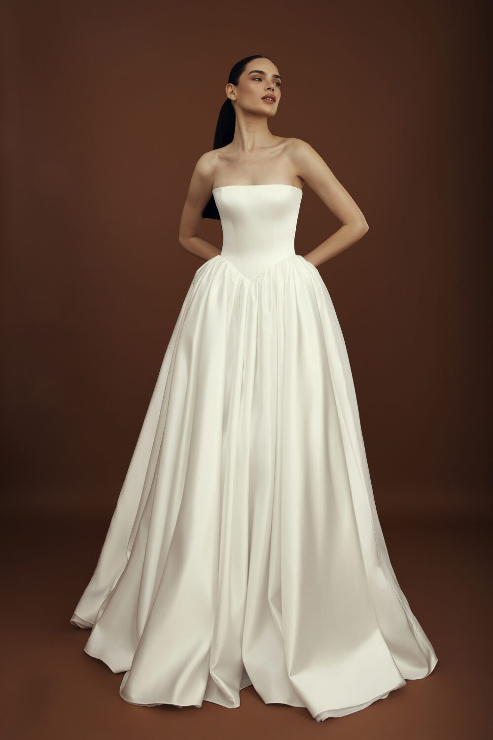 A woman models a strapless white ball gown with a structured drop-waist bodice and a full flowing skirt against a brown backdrop.