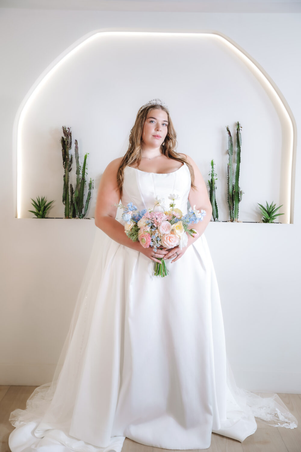 A woman wearing a tiara holds a pastel floral bouquet while posing in a white scoop-neck ball gown in front of an illuminated arch and tall cacti.