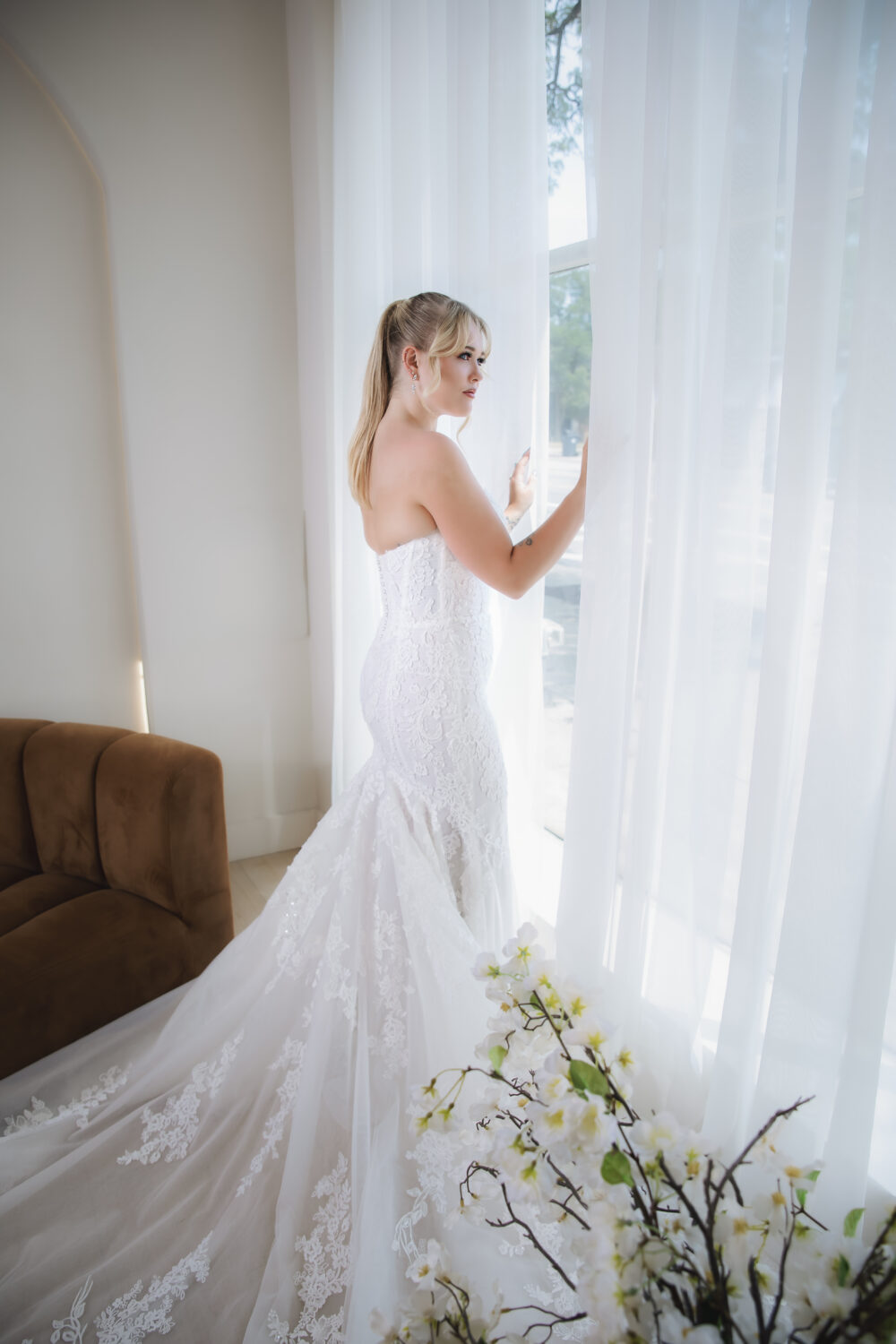 A woman in a fitted strapless lace wedding gown stands beside sheer white curtains, with her long train extending across the floor.
