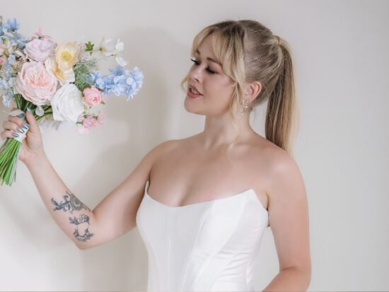 A woman in a strapless white wedding dress holds a pastel bouquet of pink, blue, and white flowers against a neutral wall.