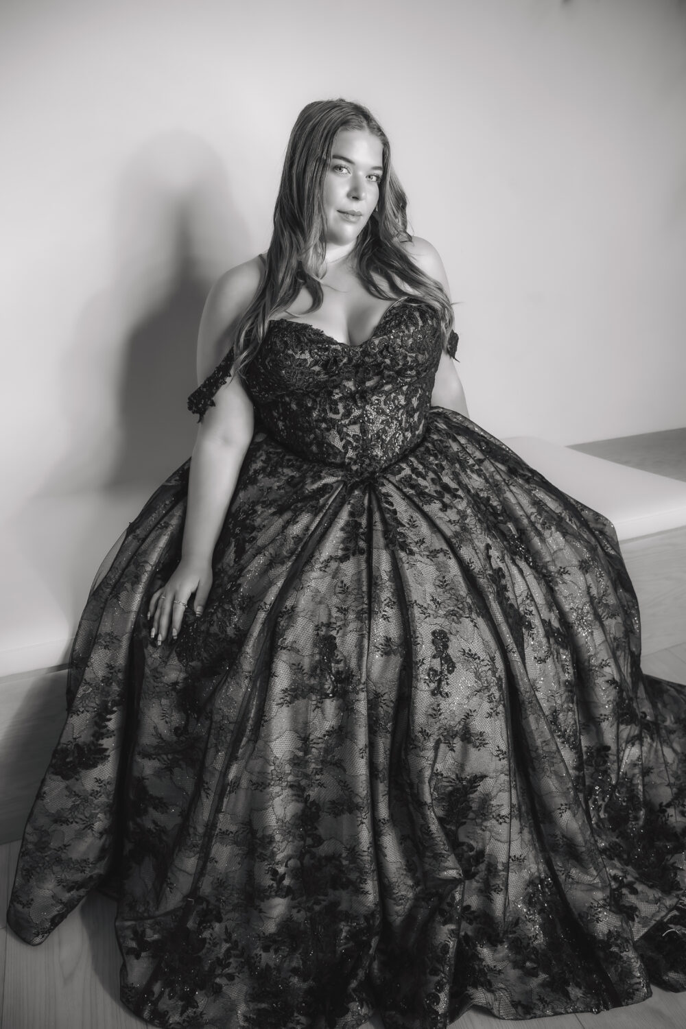 A woman poses in a black-and-white portrait wearing a dramatic dark floral ball gown with a full skirt and fitted bodice.