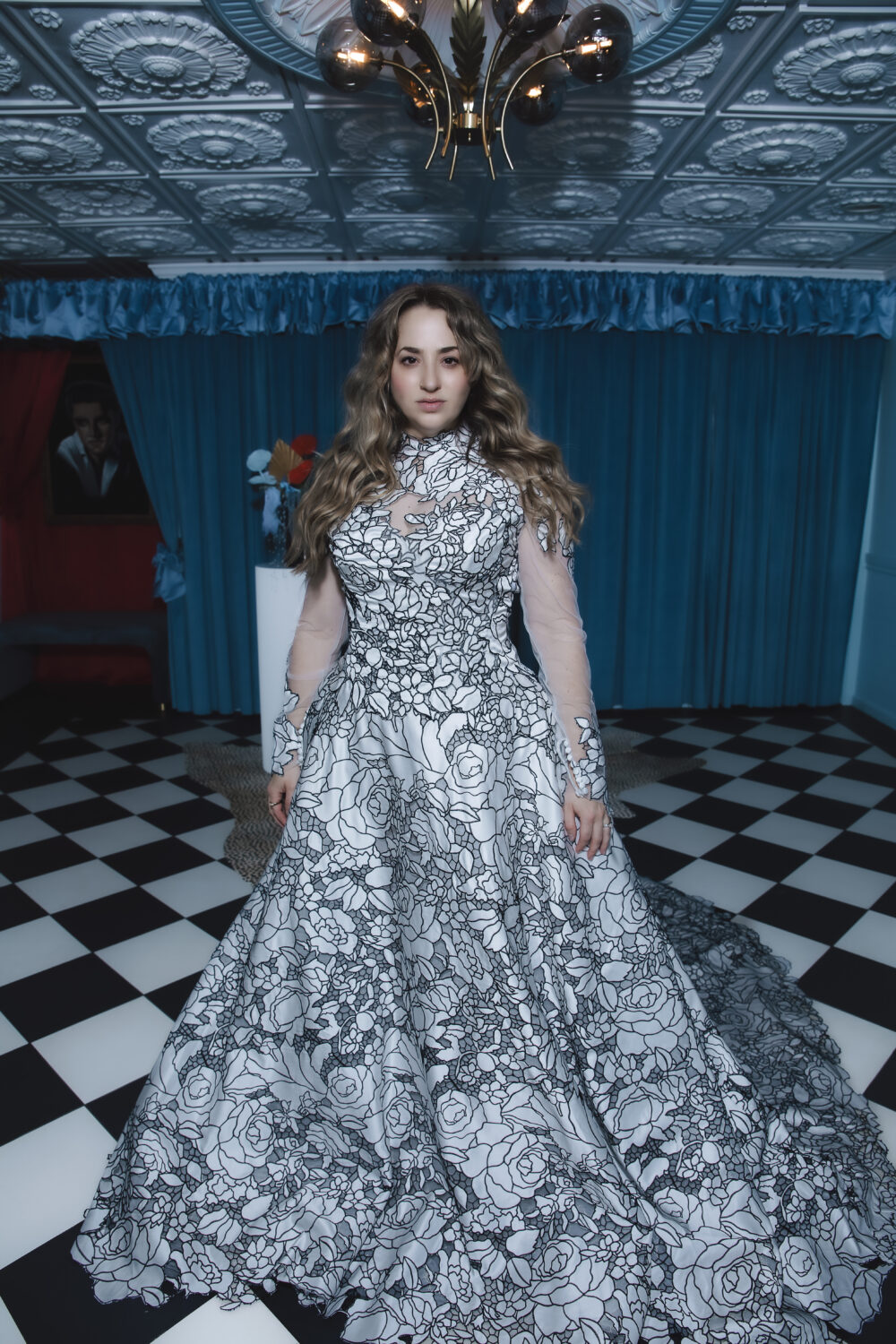 Bride in a dramatic black-and-white floral ball gown with sheer long sleeves standing in a room with blue curtains and a checkered floor.